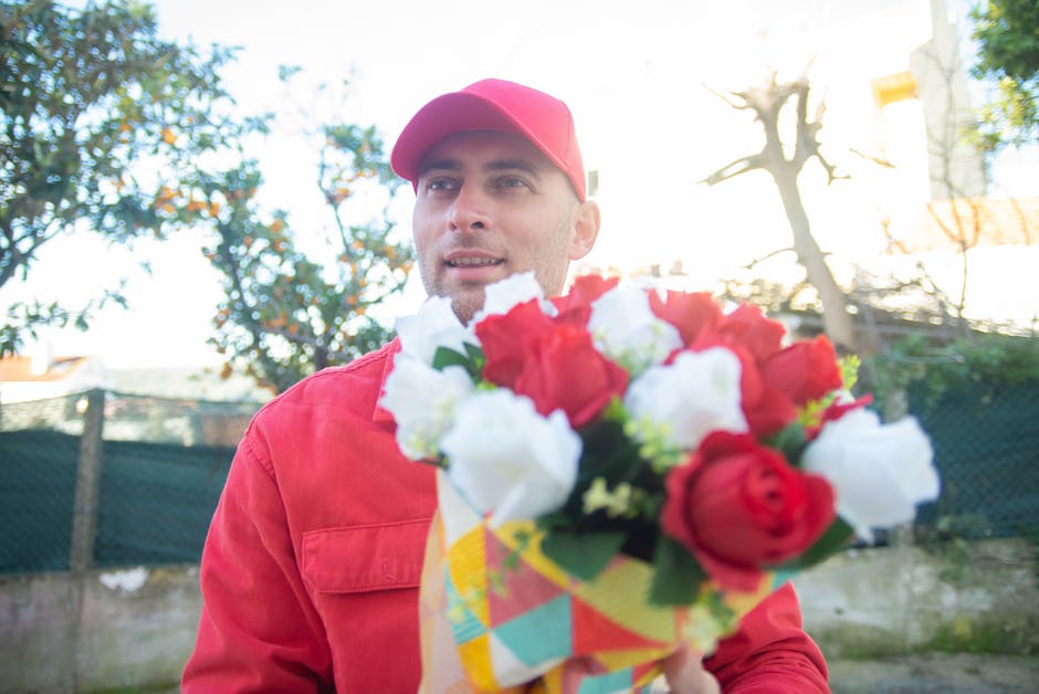 A male florist wearing a red jacket and matching red cap stands outdoors during daylight, holding a vibrant bouquet of fresh flowers wrapped in colorful paper. The bouquet features red and white roses