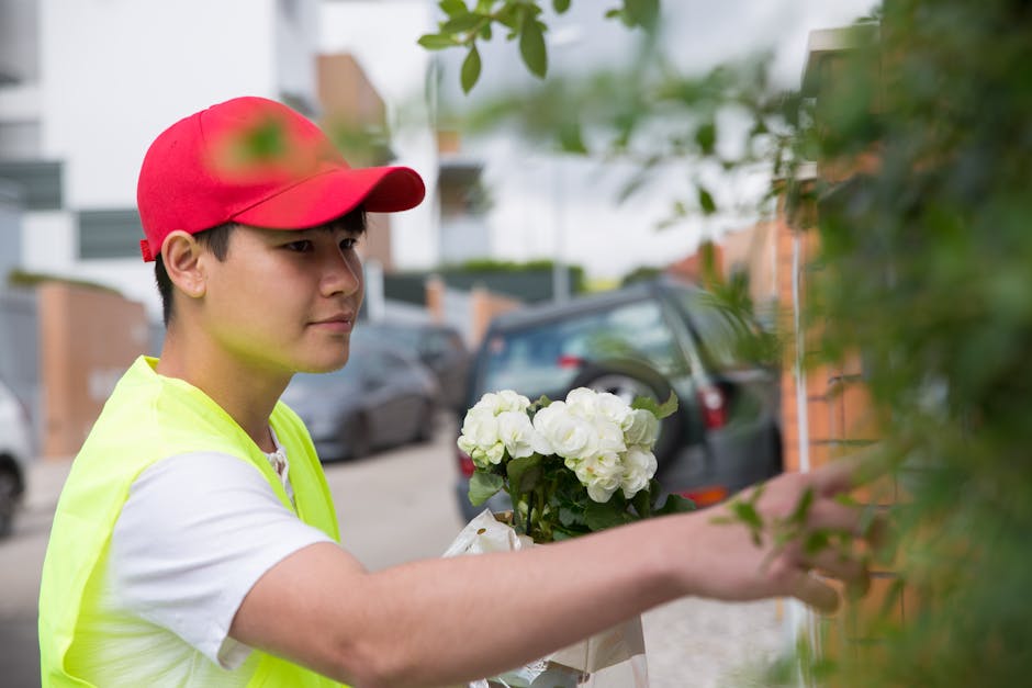 A young delivery person wearing a red cap and a neon yellow vest is carefully holding a bouquet of fresh white flowers wrapped in white paper while reaching out to deliver them, with parked cars and r